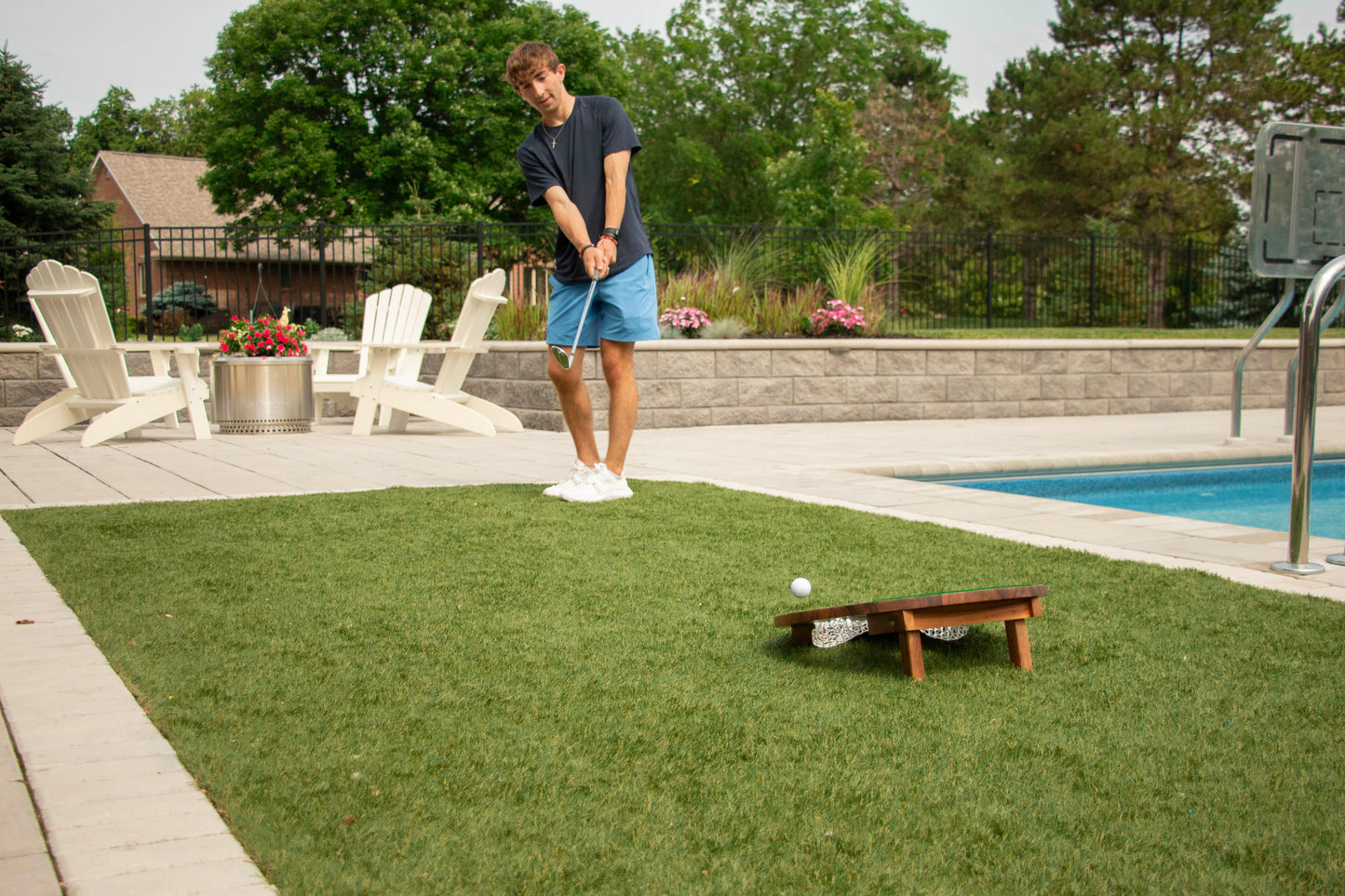 Person chipping a golf ball by a pool with green grass and trees in the background