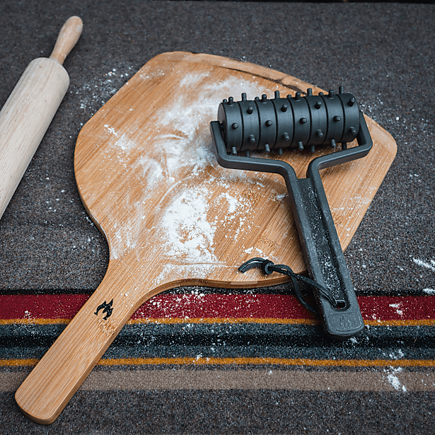 Black dough roller on a wooden pizza peel with flour on a dark surface