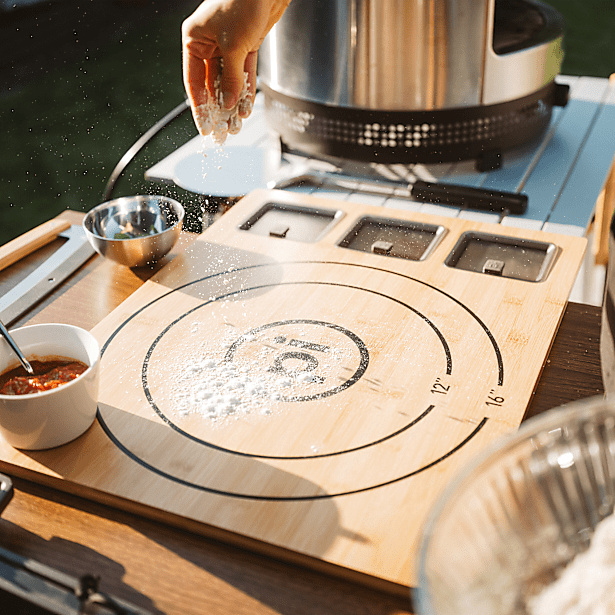 Person sprinkling flour on a wooden board with pizza measurement markings, next to a pot on a stove.