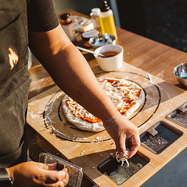 Person preparing a pizza on a wooden board with various ingredients and utensils around.